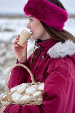 Pembe kürk bantlı ve kışlık ceketli çekici genç bir kadın dondurma külahının tadını çıkarırken sakin bir kış sahnesinin karşısında duran dondurma külahıyla dolu sepeti tutuyor..