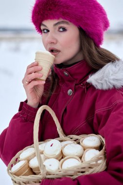 Kışın pembe kürk şapka ve kestane rengi palto giyen genç bir kadın, bir sepet dolusu dondurmayı soğuk zemine karşı tutarken tek bir dondurma külahından zevk alıyor.