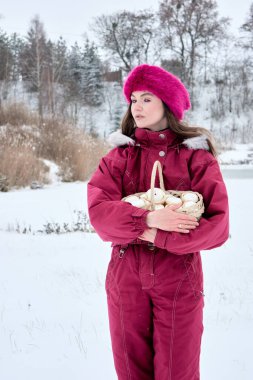 Mor kayak takımı ve kürk şapkalı güzel bir kadın, elinde beyaz paskalya yumurtalarıyla dolu bir sepet, karlı bir açık hava sahnesinde duruyor, sakin bir ifadeyle başka tarafa bakıyor..