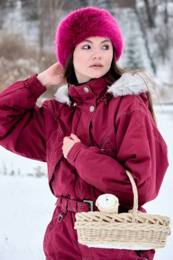 Canlı pembe kürk şapkalı ve bordo bir kış tulumuyla büyüleyici genç bir kadın dışarıda poz veriyor, karlı bir manzaranın ortasında büyüleyici bir sepet taşıyor, zarafet sergiliyor..