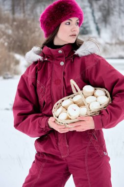 Canlı pembe bir kar elbisesi giymiş genç bir kadın ve uyumlu bir şapka zarif bir şekilde beşikte nefis dondurma külahlarıyla dolu hasır bir sepet, karlı bir kış manzarasına karşı kurulmuş..