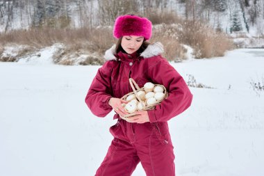 Pembe kayak takımı giymiş çekici genç bir kadın, karla kaplı bir kış manzarasında duruyor ve elinde bir sepet dolusu marşmelov tutuyor, dingin güzelliğin ve doğanın tatlı hoşgörüsünün tadını çıkarıyor.