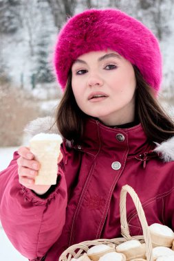 Canlı pembe kış şapkası ve paltosuyla büyüleyici genç kadın sakin, karla kaplı bir kış harikalar diyarının ortasındaki kırsal bir sepetten kar dolu waffle külahlarını sunar..