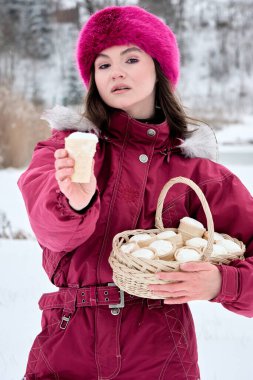 Canlı pembe şapkalı çekici genç kadın kar örtülü bir arka planda büyüleyici bir sepetten dondurma külahlarını sunuyor. Kış neşesi ve cömertliği çağrıştırıyor..