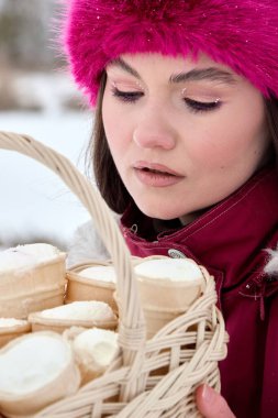 Canlı pembe şapkalı genç bir kadının büyüleyici bir yakın çekimi, dondurma külahlarıyla dolu bir sepeti nazikçe tutuyor, sakin bir kış arkaplanına dayanıyor. Neşe ve beklenti saçar. İkramlara bakar ve onları mükemmel kılar.