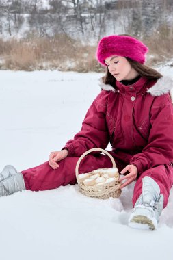 Çekici genç bir kadın, kahverengi bir kar elbisesi ve canlı pembe bir kürk şapkası ile karlı bir arazide oturuyor. Elinde lezzetli şeylerle dolu bir sepet tutuyor. Huzurlu ve peri masalı gibi cazibesiyle büyüleyici bir kış sahnesi yaratıyor..