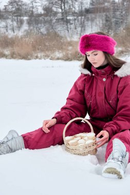 Canlı pembe bir kayak takımı giymiş genç bir kadın, düşünceli bir şekilde soğuk bir kış günü manzaralı bir yerde dondurmayla dolu bir sepete bakıyor..