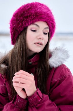 Kışlık giysiler içindeki güzel bir kadın dışarıda karda canlı pembe bir kürk şapkası ve kürk kapüşonlu sıcak bir kış ceketi giyerek ellerini nazikçe ısıtırken yakalanıyor..