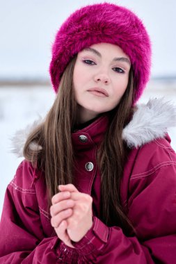 Benzersiz makyajı ve kışın açık havada poz veren mor kürklü şapkası olan çekici genç bir kadın, karlı bir ortamda kameraya bakarken kürklü kapüşonlu kırmızı bir parka giyiyor..