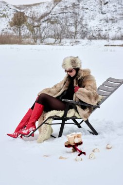 Kürk mantolu ve kırmızı çizmeli zarif bir kadın, kar üzerinde tuhaf bir dönüşle kış pikniğinin tadını çıkarıyor. Açık havada gerçeküstü bir deneyim için dökülmüş koniler ve şık aksesuarlar sunuyor..