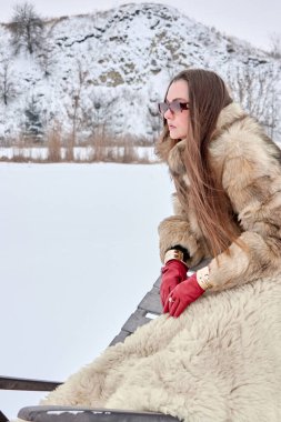 Uzun kahverengi saçlı, kürk manto, kırmızı eldiven ve güneş gözlüğü takmış zarif bir kadın, düşünceli bir şekilde kar manzarasına bakıyor, yüksek moda ve zamansız stil sergiliyor..
