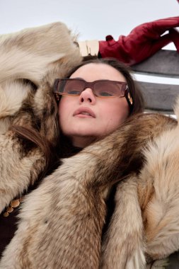 Kahverengi saçlı, şık güneş gözlüğü takan ve kırmızı eldivenli zarif esmer kadın, ahşap bir yüzey üzerinde rahat bir şekilde yatıyor, lüks bir kürk manto ile çevrili, kendine güvenini ifade ediyor..