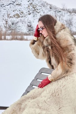 Şık kürklü ve çarpıcı kırmızı eldivenli zarif bir kadın sakin bir kış sahnesinde bir bankta oturuyor, sofistike ve soğuk hava modasının özünü karlı bir zemine yansıtıyor..