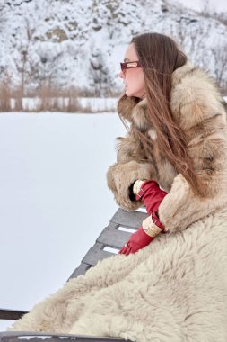 Lüks kürklü ve kırmızı deri eldivenli şık bir kadın, kar manzaralı bir bankta oturuyor, düşünceli bir bakışla mesafeye bakıyor, kış modası..