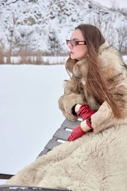 Şık kürklü ve modaya uygun güneş gözlüklü sofistike bir kadın kış manzarasının tadını çıkarıyor, zarif ve son moda bir kar manzarası sergiliyor..
