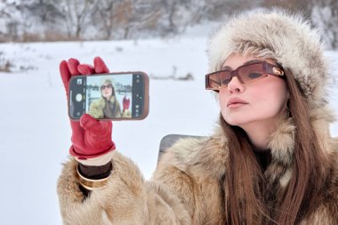Kürk manto, kürk şapka ve şık güneş gözlüğü takmış zarif bir kadın, karlı bir arkaplanda selfie çekiyor. Modaya uygun kış tarzını, klasik bir havası ve kendine güvenen bir ifadeyle sergiliyor..