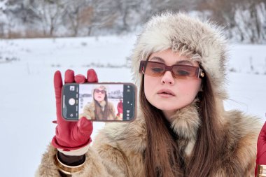 Kürk şapkalı ve güneş gözlüklü şık bir kadın karlı bir ortamda selfie çekiyor, kış stili, teknoloji ve modern kendini ifade ediyor, mevsimlik ve yaşam tarzı için mükemmel..
