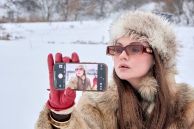 Kürk manto ve şapka giymiş güzel bir esmer kadın kış manzarasında kendi portresini çekiyor, akıllı telefonunu kullanarak, moda ve teknolojinin bir karışımını sergiliyor..