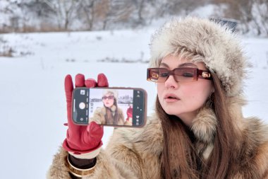 Kürk şapkalı ve paltolu şık bir kadın, kar manzarasında kışın keyfini çıkarırken akıllı telefonuyla modayı yakalamak için bir selfie çekiyor.