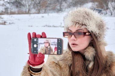Kürk şapka ve paltosu da dahil olmak üzere kışın giyinmiş şık bir kadın, akıllı telefonunu karlı bir açık hava ortamında selfie çekmek için kullanıyor. Mevsimlik kıyafetleri modern teknolojiyle harmanlıyor..