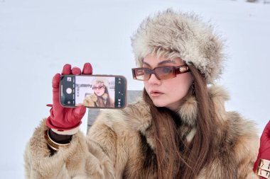 Kürk şapkalı ve ceketli göz kamaştırıcı bir kadın şık telefonuyla özçekim anı yakalıyor. Kaliteli güneş gözlüklerini ve canlı kırmızı eldivenlerini açık havada el değmemiş, karlı bir kış sahnesine vurguluyor. Zarafet ve stil katıyor..