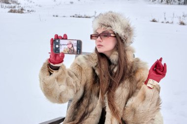 Şık bir kadın kış harikalar diyarında, zarif bir kürk ve şapka giyip, klasik modayla sosyal medya için çağdaş mobil teknolojiyi harmanlayan bir selfie çekiyor..