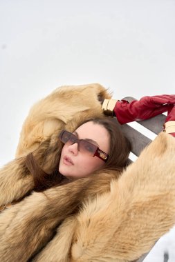 Kürk ceketli ve güneş gözlüklü zarif genç kadın, karlı bir günde dışarıda kırmızı deri eldivenler ve altın bileziklerle poz veriyor, lüks ve moda anlayışı sergiliyor..