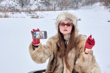 Kürk şapkalı ve kürklü çekici bir kadın karda akıllı telefonuyla selfie çekiyor. Güneş gözlüklü ve eldivenli şık bir kız. Kış selfie konsepti..