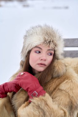 Kürk şapkalı ve ceketli çekici bir kadın kış boyunca parkta bir bankta otururken mesafeye bakıyor, kış modasını ve açık havada poz veren dış görünüşü somutlaştırıyor.