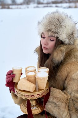 Kürk şapkalı ve ceketli güzel bir kadın zarif bir şekilde sunar. Karlı kış manzarasının ortasında içi dondurma külahlarıyla dolu altın bir tezgah. Lüks bir hoşgörü ve cazibe hissi uyandırıyor..