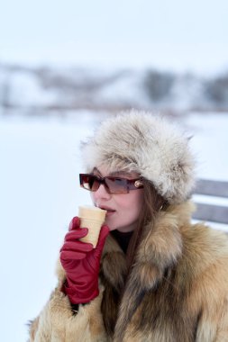 Kürk şapkalı ve şık kürklü zarif bir kadın kış manzarasında tatlı bir dondurma külahının tadını çıkarıyor. Güneş gözlüğü ve canlı kırmızı eldivenlerle tamamlanıyor.