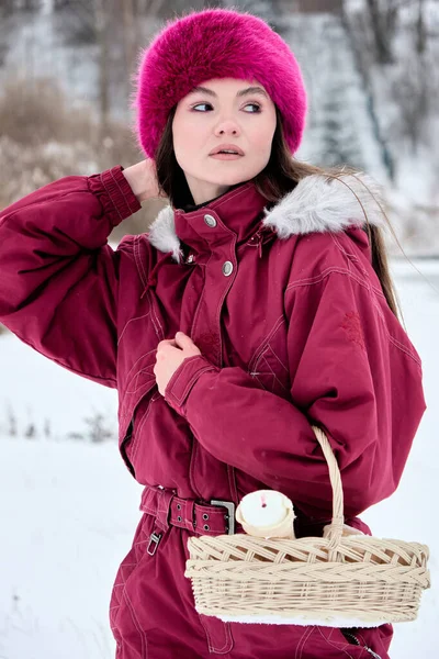 Mor renkli bir kayak takımı ve uyumlu kürklü bir şapkası olan çekici genç bir kadın elinde hasır bir sepet ve beyaz bir eşyayla açık havada güzel bir kış sahnesinde poz veriyor..