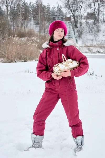 Karlı bir kış sahnesinde, macenta bir kar elbisesi giymiş bir kadın, doğada tuhaf ve mevsimsel bir an gösteren bir sepet dolusu marşmelovla poz verir..