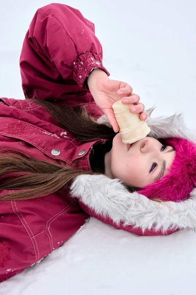 Kışlık giysiler içinde genç bir kadın, karların içinde uzanmış, ağzına bir gözleme fincanı tutarak, güzel kış manzarasının ve doğanın huzurunun tadını çıkarırken, tatlı bir ziyafetin tadını çıkarıyor..