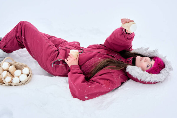 Delighted young woman clad in winter apparel relishes ice cream in the snow, lying amidst a whimsical winter scene and a basket of cones, enjoying the delightful moment with pleasure.