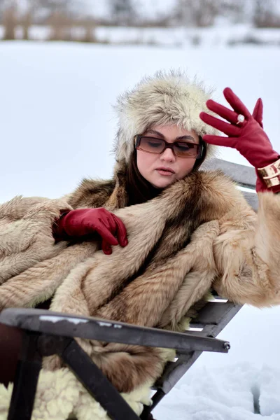 Şık bir kürk manto ve şapkayla süslenmiş entelektüel bir bayan, kar yağışlı bir bankta poz veriyor, şık kırmızı deri eldivenlerini ve zarif mücevherlerini sergiliyor, kendine güvenini sergiliyor.