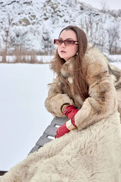 Kürklü ve şık güneş gözlüklü zarif bir kadın, kış günü, karlı bir zemine karşı lüks ve modern modanın bir karışımını sergileyerek güvenli bir şekilde dışarıda poz veriyor..