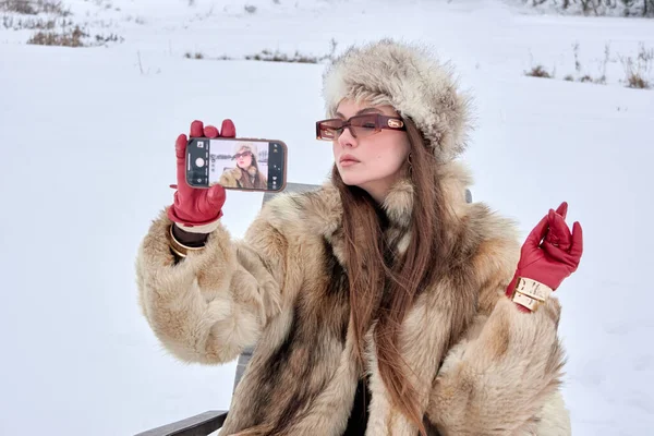 Şık bir kadın kış harikalar diyarında, zarif bir kürk ve şapka giyip, klasik modayla sosyal medya için çağdaş mobil teknolojiyi harmanlayan bir selfie çekiyor..