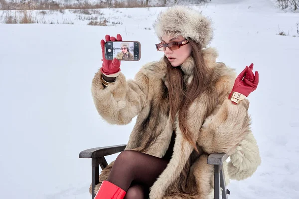Kürk mantolu ve şapkalı çekici bir kadın, kar manzaralı bir ortamda akıllı bir telefonla selfie çekiyor. Kış zarafetini ve modern teknolojiyi gözler önüne seriyor. Kameraya poz veriyor..