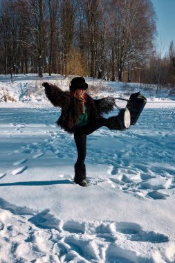 Sakin bir kış manzarasının ortasında, kürk manto, şapka ve eşarp giymiş bir kadın bir çanta tutarken neşe içinde dengede duruyor, parlak bir güneşin altında, karlı bir ortamda tuhaf bir manzara yaratıyor..