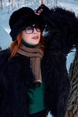 Şık kızıl saçlı kadın, kış modasına uygun giyiniyor. Lüks bir kürk manto, kürk şapka, şık bir güneş gözlüğü ve zarif bir eşarp. Karla kaplı bir orman zeminine karşı sofistike bir dokunuşla poz veriyor..