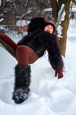 Kış modasına dalmış zarif bir kadının büyüleyici bir görüntüsü. Karla kaplı bir hamakta, kürk, şapka ve çizme giymiş bir şekilde zarif bir şekilde uzanmış..