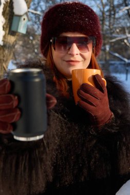 Şık bir kadın kış gününün tadını çıkarıyor sıcak ve soğuk içeceklerle, kürk şapkası ve ceketiyle poz veriyor, elinde turuncu bir kupa ve siyah bir teneke kutu tutuyor, mevsimsel karşıtlığı kucaklıyor..