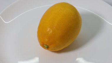 Canlı sarı bir limonu yakından çekerek, beyaz bir tabakta yavaşça dönerek turunçgillerin ten dokusunu ve pürüzsüz ışıklandırma koşulları altında parlak rengini detaylı bir şekilde gösteriyor..
