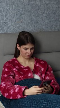 Pijamalı çekici genç bir kadın akıllı telefonuyla yatakta dinleniyor, sabahları esniyor, mutlu ve ferah bir şekilde evde uyanıyor, tembel ve rahat bir sabah geçiriyor.