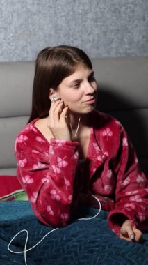 Yonca desenli rahat bir pijama giymiş, yumuşak bir battaniyeyle yatağında rahatça oturan, kulaklıklarıyla müzik dinleyen ve akıllı telefon uygulamasını kullanan çekici bir genç kadın..