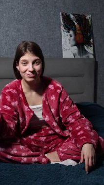 Moda bir pijama seti içinde mutlu bir genç kız yatağında rahat bir şekilde oturur ve etkin bir şekilde görüşlerini güven ve coşkuyla paylaşır..