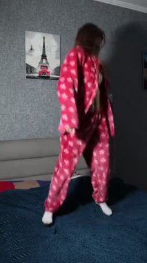 Enerjik ve kaygısız, canlı pembe pijamalı genç bir kadın bir yatakta zıplar ve dans eder, neşe saçar ve neşeli bir anda pozitif titreşimler yayar..