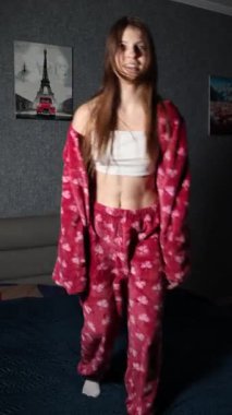 Enerjik genç bir kadın, yoncalarla dolu rahat kırmızı pijamalar giyiyor, mutlu bir şekilde yatağına atlıyor, sonra yere düşüyor ve yatak odasında, parmaklarıyla barış işaretleri gösteriyor.