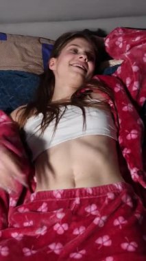 Çiçek desenli kırmızı pijama seti içinde uzun saçlı mutlu bir kız yatakta dinleniyor, karnını gösteriyor, geniş bir gülümseme ve rahat bir yatak odasında barış işaretleri gösteriyor.